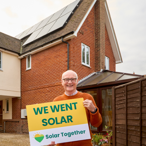 Man stands in front of solar panels on his house, holding up a sign which reads: We Went Solar. Solar Together. 