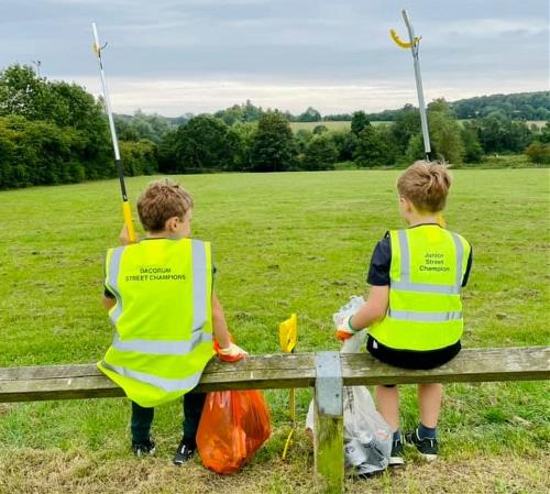 Dacorum Junior Street Champions Two boys sat on a fence, facing away from the camera with their litter pickers in the air. They wear hi-vis vests with 'Junior Street Champions' on the back.